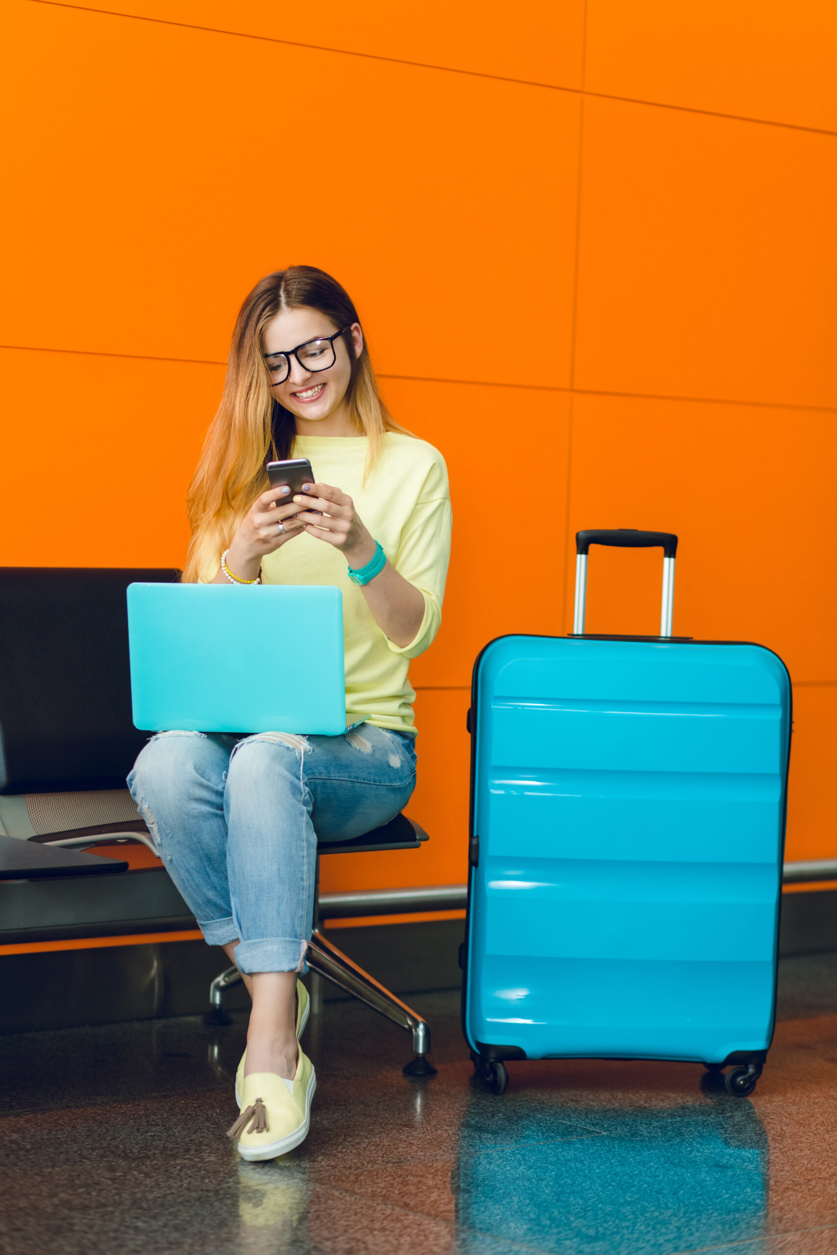 Home young girl in yellow sweater and jeans is sitting on chair on orange background. she has blue laptop on knees and blue suitcase near. she is typing on phone